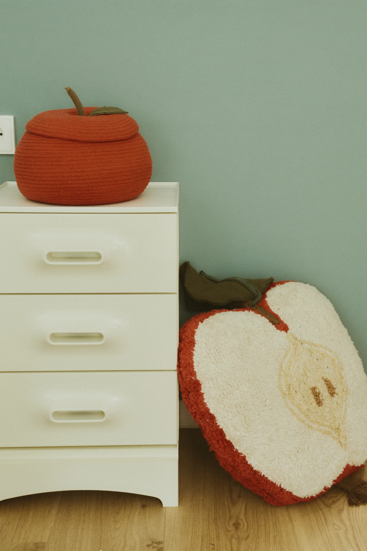 A styled interior scene showing a white three-drawer nightstand against a muted sage green wall, with a knitted apple storage basket in red and a large apple-half floor cushion leaning beside it.
