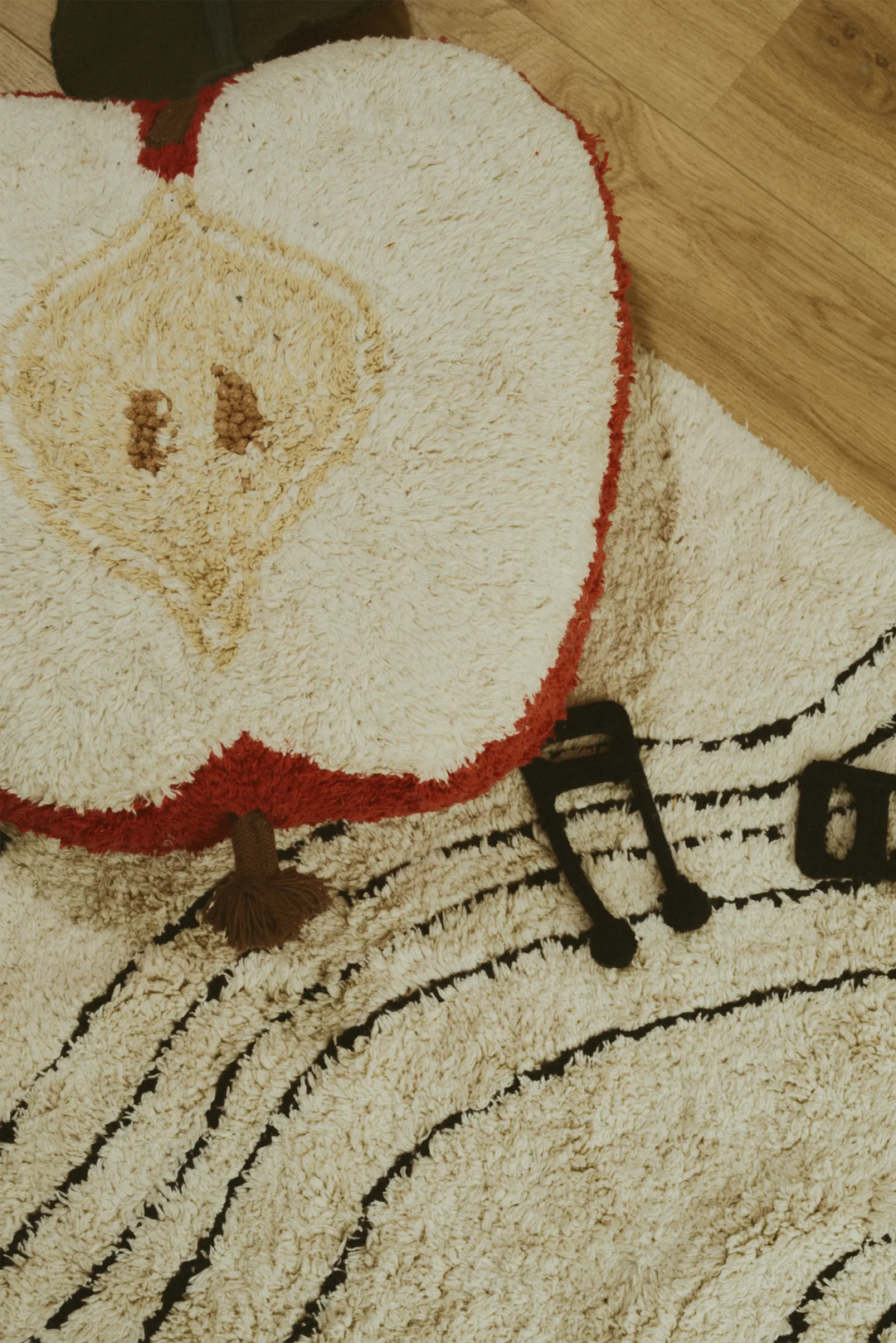 A close-up of a plush shag rug featuring a large cream-colored apple slice design with red edges and a tan seed detail, alongside black musical note and staff line patterns on a light wood floor.
