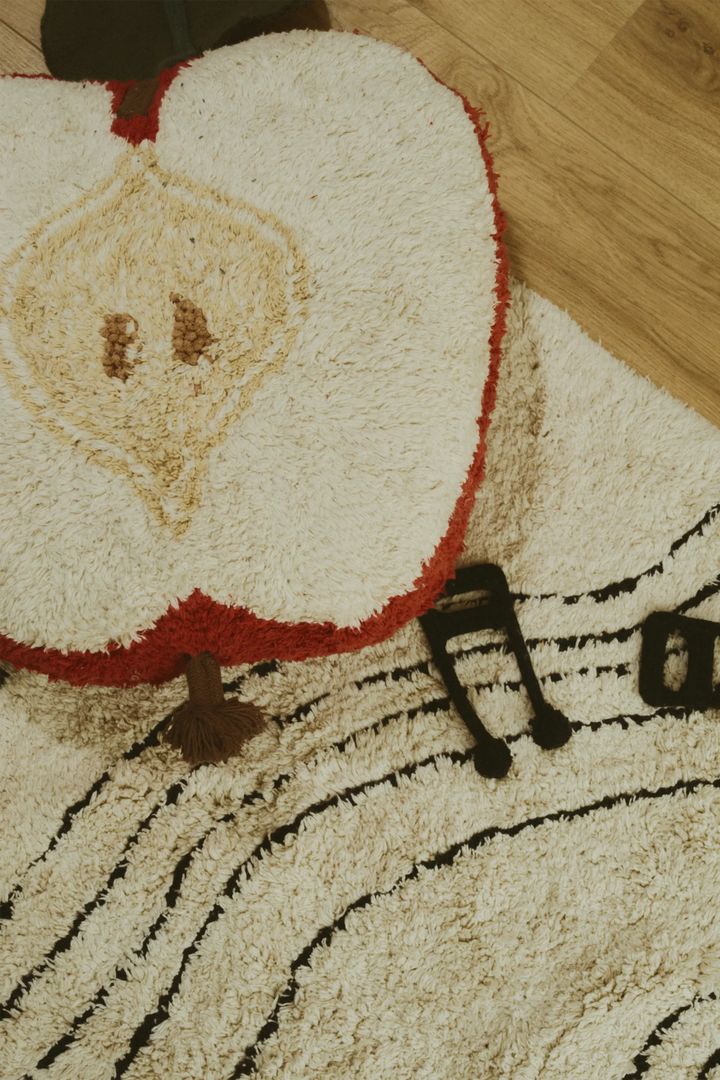 A close-up of a plush shag rug featuring a large cream-colored apple slice design with red edges and a tan seed detail, alongside black musical note and staff line patterns on a light wood floor.