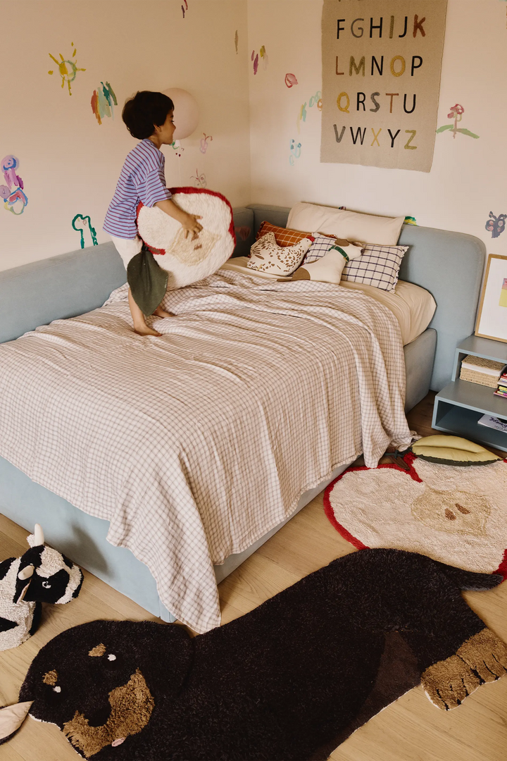 A child stands on a light blue bed in a cozy, decorated children's bedroom, holding a round white plush pillow with red trim. The room features a light wood floor, a black dog-shaped rug, a patterned sheep rug, and a wall poster showing the alphabet.