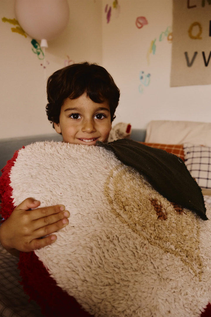A smiling young boy with dark hair holds a large, plush, textured red apple shaped floor cushion in a cozy bedroom setting.