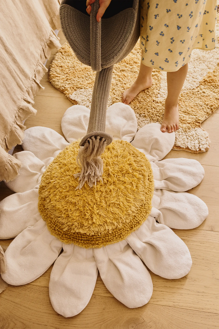 A person holds a woven straw hat over a large daisy-shaped rug with cream petals and a mustard yellow center, placed on a light wood floor.