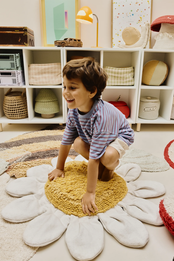 A young boy with brown hair crouches on a round, yellow knitted rug surrounded by a large white flower-shaped rug in a cozy, styled children's room.
