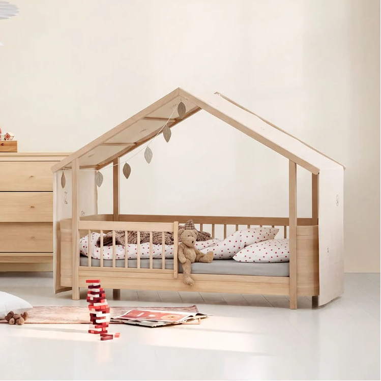 A small house-shaped wooden children's bed in oak by Oliver Furniture with a slatted guard rail and a pitched roof, styled in a bright nursery with bedding, a teddy bear, and toys on the floor.