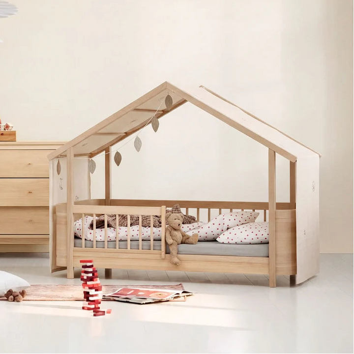 A small house-shaped wooden children's bed in oak by Oliver Furniture with a slatted guard rail and a pitched roof, styled in a bright nursery with bedding, a teddy bear, and toys on the floor.