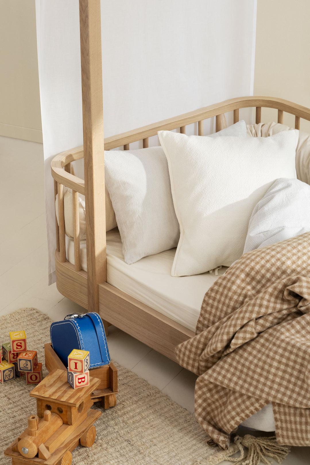 A light natural wood canopy bed with white pillows and bedding, styled in a child's bedroom with a brown gingham blanket, toys, and a blue suitcase on a woven rug.