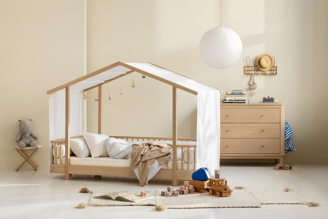 A light natural wood house-shaped canopy bed with white fabric curtains and bedding, styled in a warm, minimalist children's bedroom with a wooden dresser, white globe lamp, and toys on a rug.