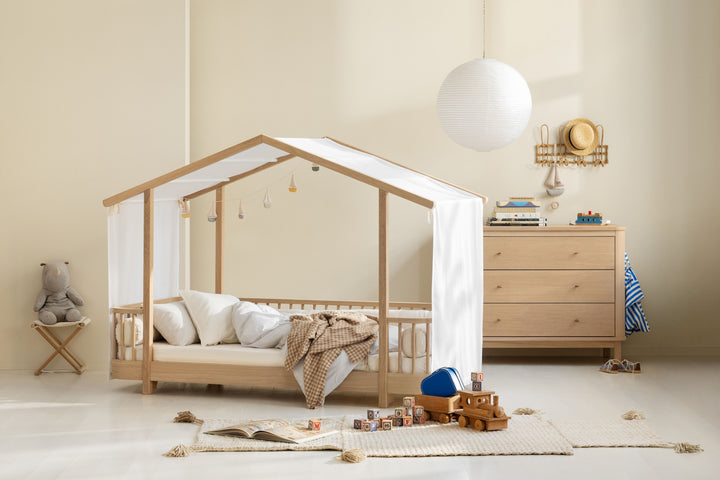 A light natural wood house-shaped canopy bed with white fabric curtains and bedding, styled in a warm, minimalist children's bedroom with a wooden dresser, white globe lamp, and toys on a rug.