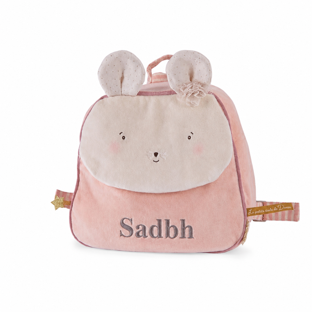 A pink plush children's backpack shaped like a mouse, featuring a cream-colored face on the front, embroidered eyes and nose, pink blush cheeks, and the name 'Sadbh' embroidered in gray. It has bunny ears on top, a top handle, and a small decorative tag on the side.