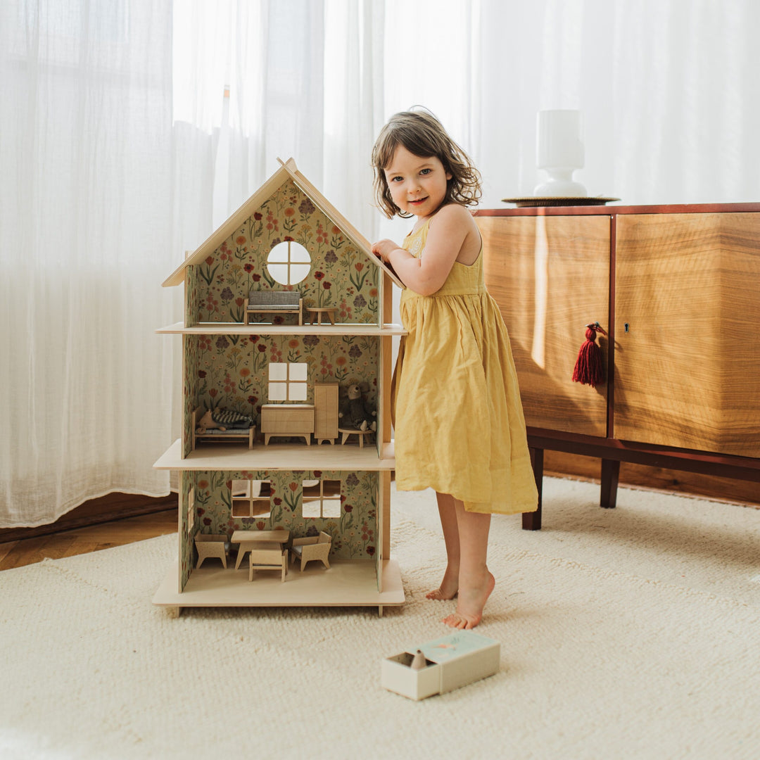 Child in a yellow dress standing next to a wooden dollhouse in a room with light-colored walls and furniture.