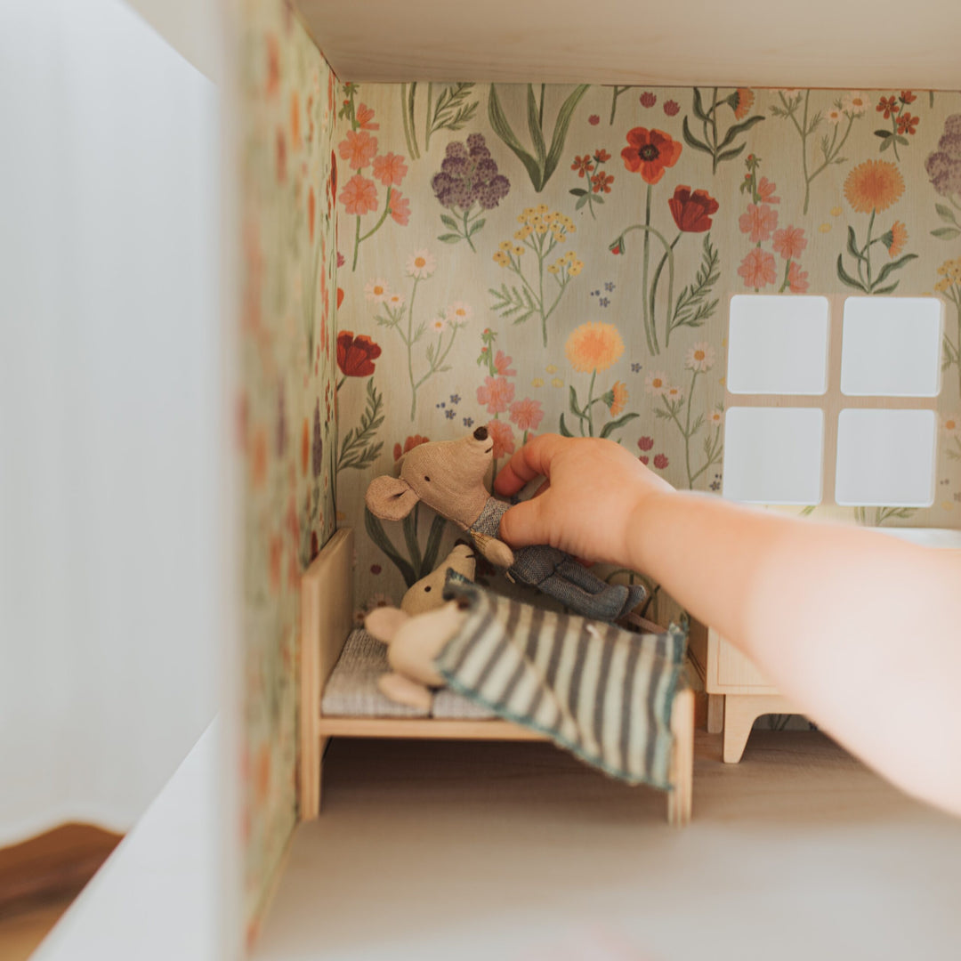 Hand placing a Maileg mouse on a bed in a dollhouse with floral wallpaper.